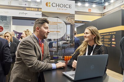 Eine Frau und ein Mann unterhalten sich lächelnd an einem Messestand von Choice Hotels. Vor ihnen steht ein Laptop, im Hintergrund sind weitere Besucherinnen und Besucher sowie Werbegrafiken des Unternehmens zu sehen.
