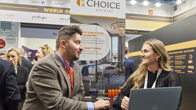 A woman and a man are smiling and talking at a Choice Hotels trade fair booth. A laptop sits in front of them, with other attendees and company promotional displays visible in the background.