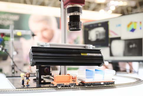 Model railway with orange locomotive and freight car passes under an optoelectronic inspection system with camera and lighting unit.
