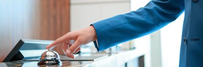 A hand in a blue suit presses a bell on a marble reception desk fitted with several computer screens.