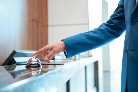 Eine Hand im blauen Anzug drückt auf eine Klingel an einem Empfangstresen aus Marmor und mit mehreren Computerbildschirmen.