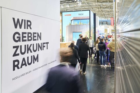 Messebesucher, die durch die Messehallen laufen und an einem Stand mit Aufschrift 