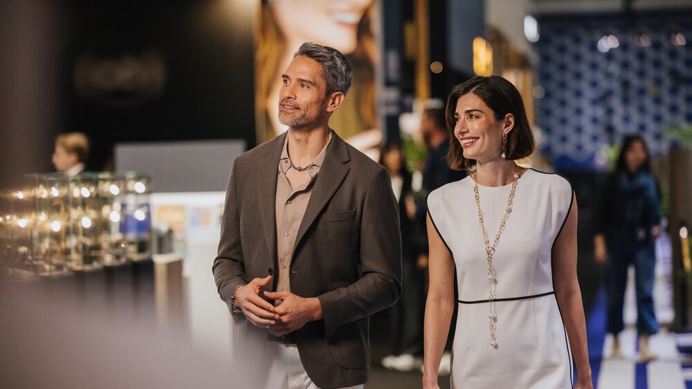 Ein Mann im grauen Blazer und eine Frau im weißen Kleid gehen in einem Raum einer Veranstaltung oder Ausstellung spazieren.