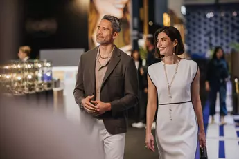 A man in a gray blazer and a woman in a white dress are walking in a room at an event or exhibition.