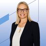 Portrait of a woman with shoulder‑length blonde hair wearing a dark blue blazer and white blouse with a small keyhole neckline in front of a light background with blue graphic lines  