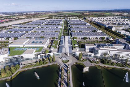Luftaufnahme des Messe München Geländes auf den Westeingang und die Hallen. Im Vordergrund ist der Messesee zu sehen.