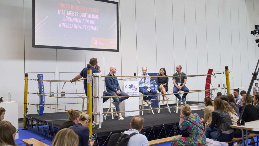 Six people are sitting on chairs around a podium with the digitalBAU logo and the title “conference & networking” in a boxing ring set up as a stage in front of the audience.