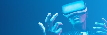 Blau getöntes Bild, auf dem ein Mann mit VR-Brille und ausgestreckten Händen vor blauem Hintergrund zu sehen ist.
