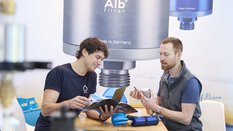 Two people are having a sales talk about water filters at a trade fair stand. An advertising poster for the water filter manufacturer can be seen in the background.