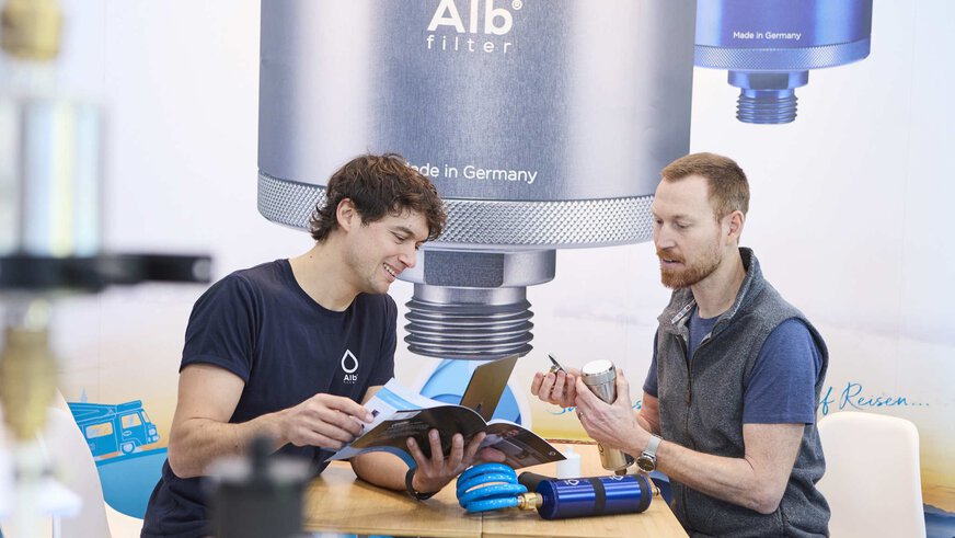 Two people are having a sales talk about water filters at a trade fair stand. An advertising poster for the water filter manufacturer can be seen in the background.