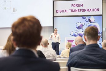The camera looks over the heads of the audience onto a stage on which a person is presenting something. 