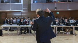 Ein Redner im Anzug von hinten, der zu einem interessierten Publikum spricht. In einem verglasten Raum oben rechts sitzt die Technikregie.