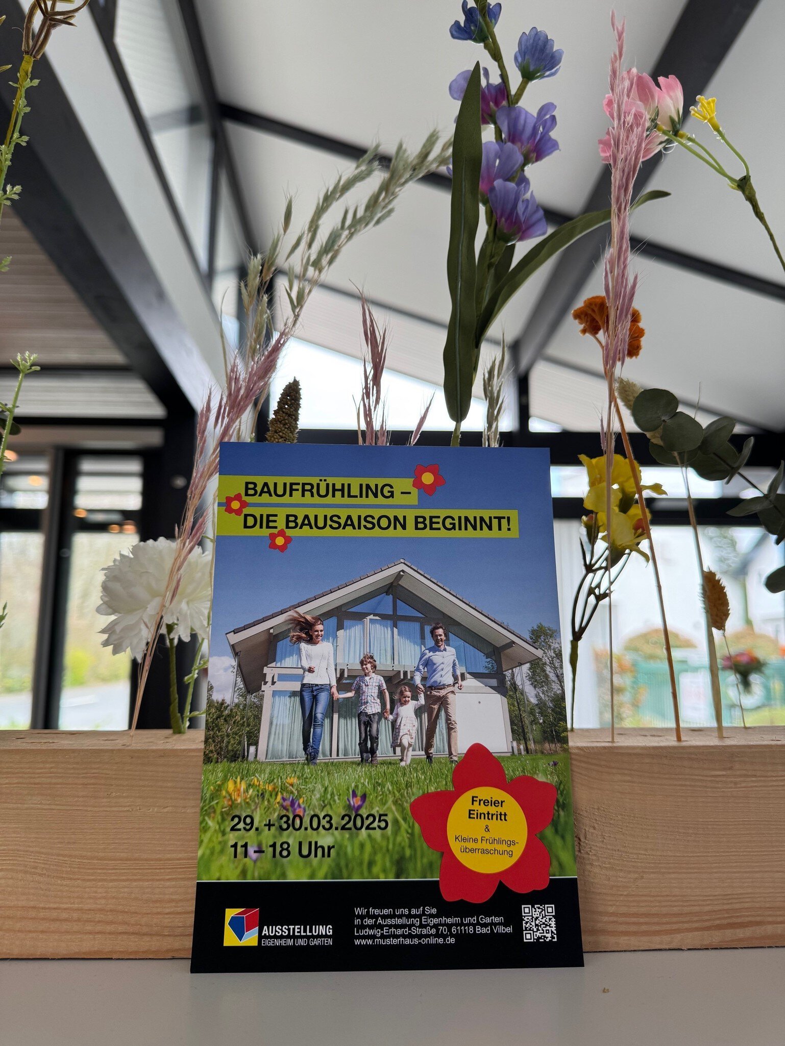 Flyer mit Informationen zur Veranstaltung „Baufrühling 2025“, lehnt an einem Holzblock, in dem ein paar lange Blumen stecken.