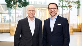Zwei Männer in Anzügen und mit Brillen, einer mit dunklen Haaren und Bart und einer mit Glatze, stehen vor einer hellen Fensterfront und lächeln.