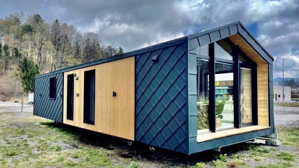 Modernes kleines Haus mit Holz- und dunkler Metallverkleidung, gelegen in einem offenen Bereich mit bewölktem Himmel und Bäumen im Hintergrund.