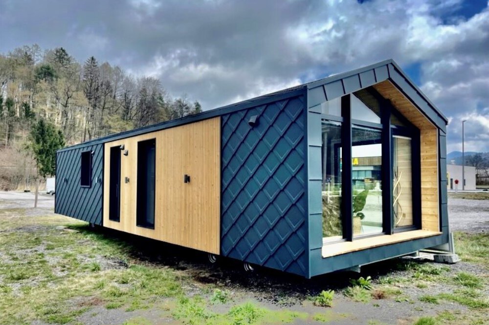 Modernes kleines Haus mit Holz- und dunkler Metallverkleidung, gelegen in einem offenen Bereich mit bewölktem Himmel und Bäumen im Hintergrund.