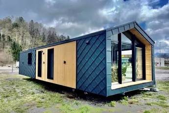 Modernes kleines Haus mit Holz- und dunkler Metallverkleidung, gelegen in einem offenen Bereich mit bewölktem Himmel und Bäumen im Hintergrund.