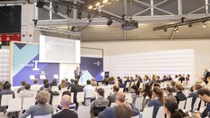 A speaker speaks on the munich_i stage in front of a full audience.