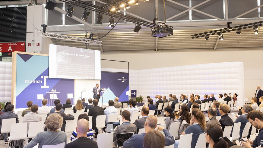 A speaker speaks on the munich_i stage in front of a full audience.