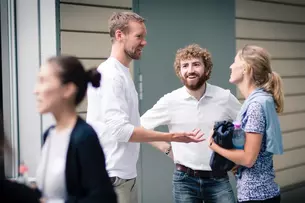 Drei Personen stehen draußen und unterhalten sich angeregt, während andere im Vordergrund vorbeilaufen.