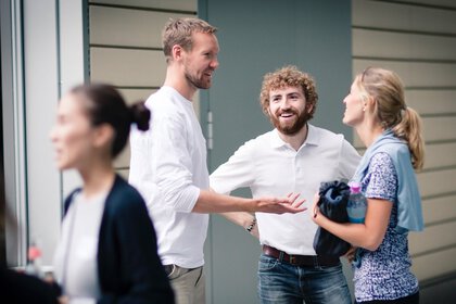 Drei Personen stehen draußen und unterhalten sich angeregt, während andere im Vordergrund vorbeilaufen.