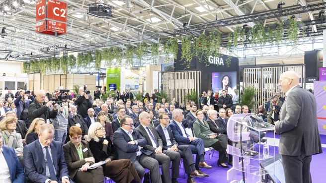 A speaker speaks from a podium in Hall C2 at BAU, in front of a large seated audience and several members of the press.