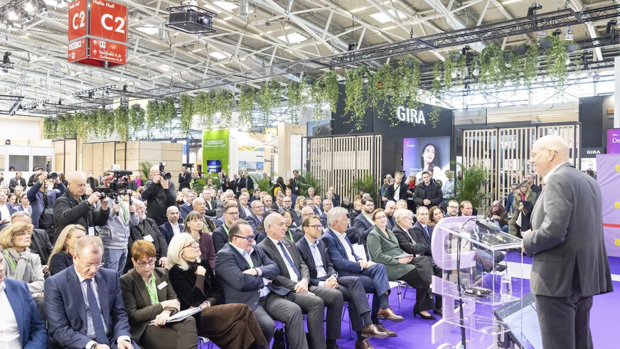Auf der BAU steht ein Redner an einem Podium vor einem großen sitzenden Publikum und mehreren Presseleuten.