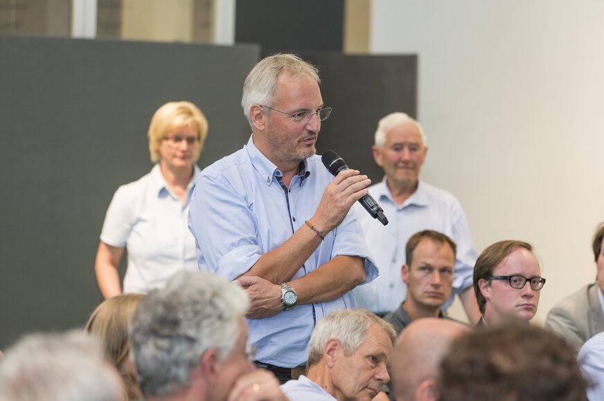 A man in a blue shirt and glasses holds a microphone in his hand as he stands among a seated audience.