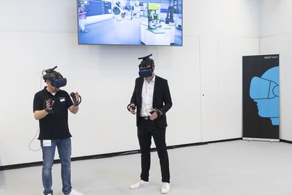 The picture shows two people wearing virtual reality headsets and holding controllers. They are standing in a room with screens displaying VR content.