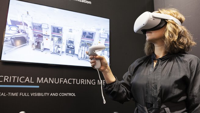 Frau mit weißem VR-Headset und Controller in der Hand steht an einem Stand auf der productronica. Hinter ihr auf dem Bildschirm wird das Bild des VR-Headsets angezeigt.