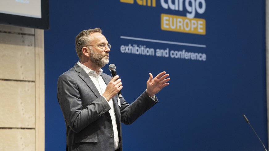 A man speaks into a microphone on a stage. Behind him, the logo of the 