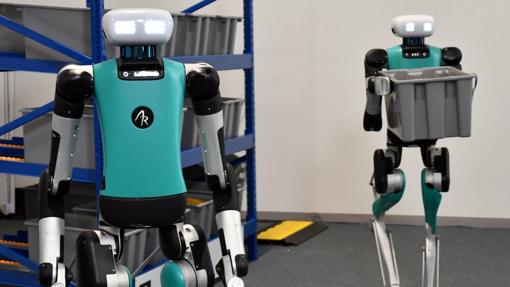 Zwei humanoide Roboter in einem Lagerhaus, einer trägt einen grauen Behälter, im Hintergrund sind Regale zu sehen.