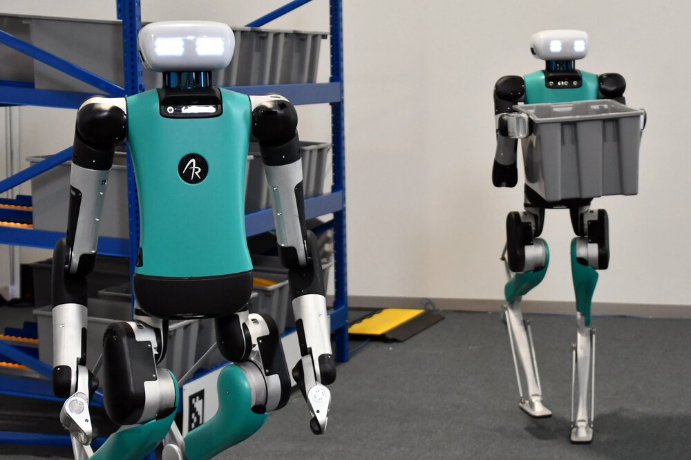 Two humanoid robots in a warehouse, one carrying a grey container, shelves can be seen in the background.