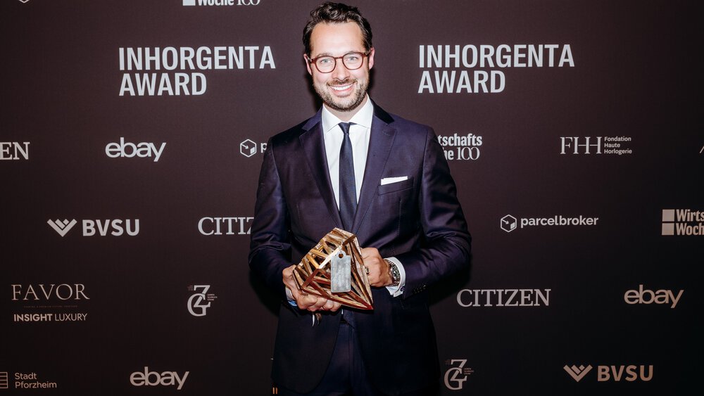 A smiling man with dark hair, a beard, glasses and wearing a suit stands in front of a photo wall of the INHORGENTA AWARD and holds a trophy.