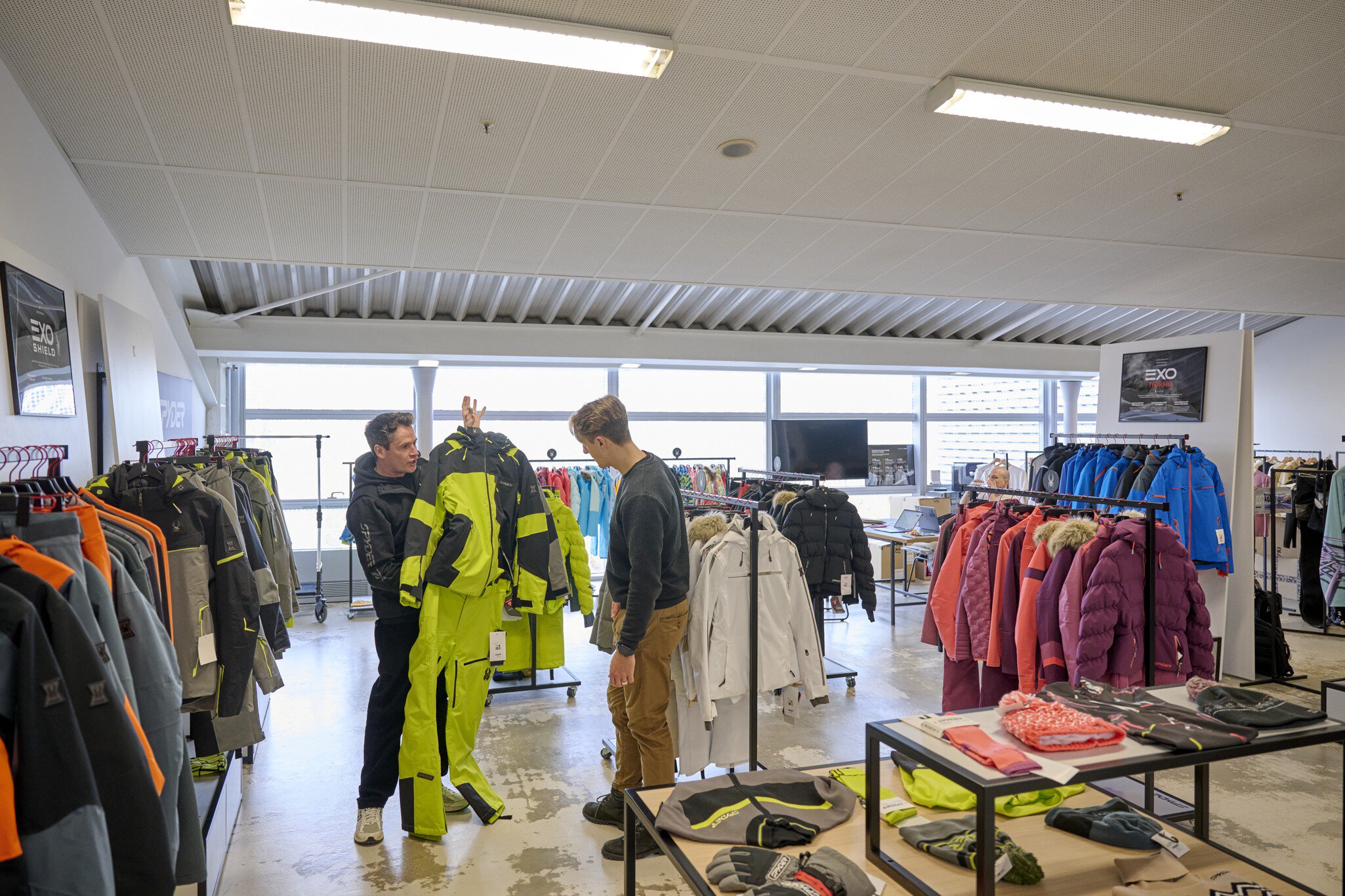 Eine Person zeigt einer anderen einen Ski-Anzug auf der Ausstellungsfläche des MOC Ordercenters. Drum herum sind viele volle Kleiderständer zu sehen.