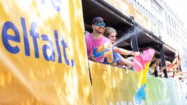At a sunny outdoor event, people on a parade float are splashing water and waving colorful flags.