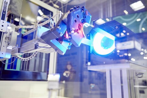 A robotic arm operates near a bright blue light at a technology exhibition, with blurred figures in the background.