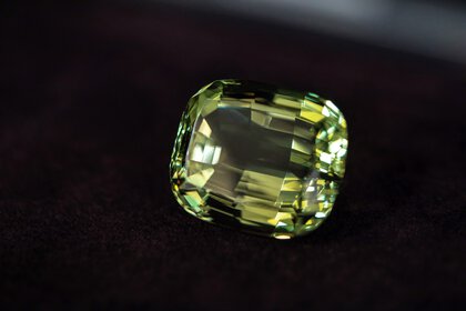 A large, faceted green gemstone resting on a dark, textured surface.
