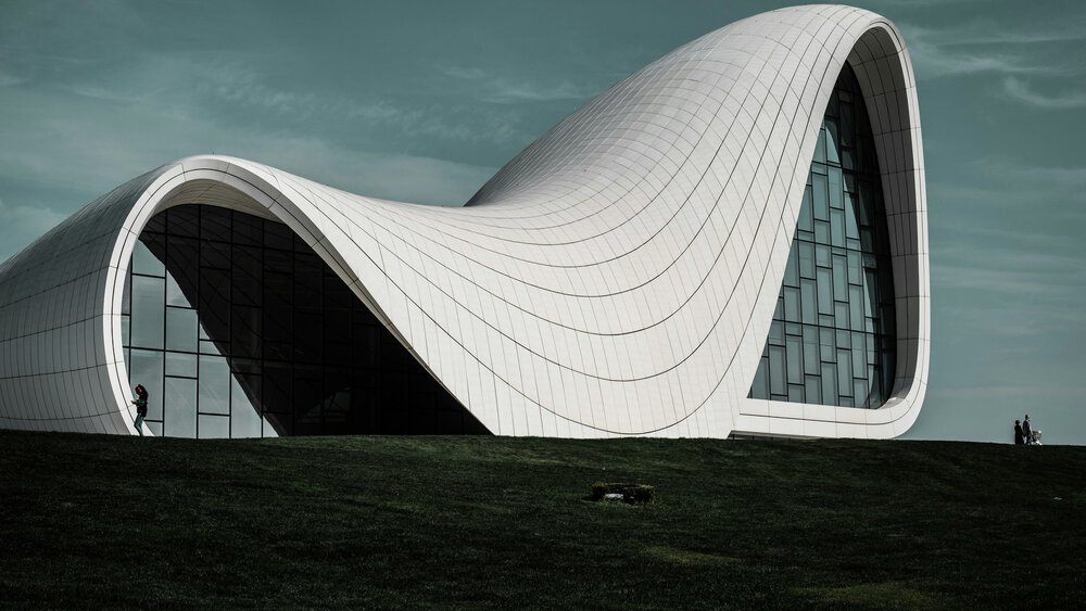 Futuristic white building with flowing curves and glass, on green grass under a cloudy sky.