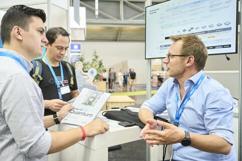 Beratungsgespräch an einem Messestand der World of Quantum mit Präsentation einer Quantencomputing-Roadmap auf einem Bildschirm.