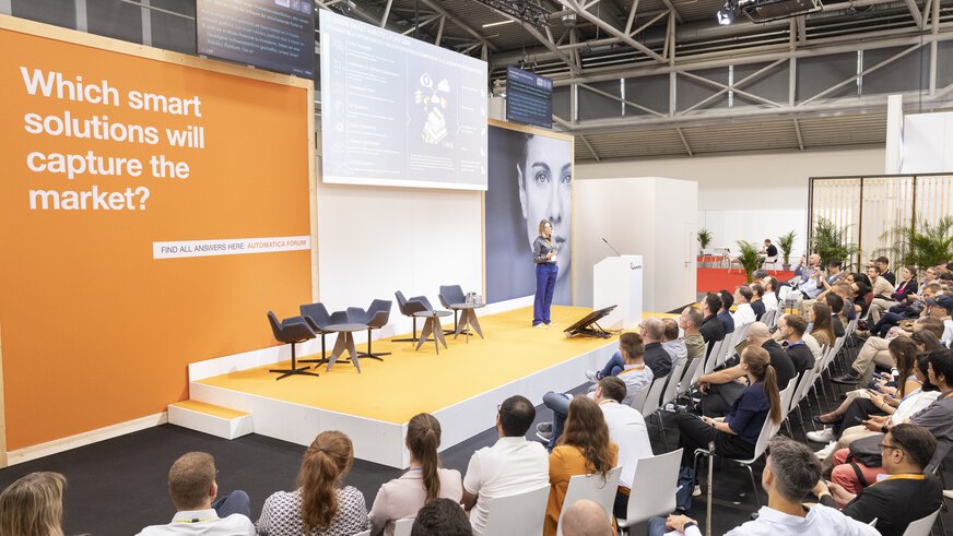 Eine Rednerin präsentiert auf einer großen Bühne des automatica Forums vor einem voll besetzten Publikum. Hinter ihr ist eine große Leinwand mit einer Präsentation zur Smart Robotics Plattform zu sehen. Links im Bild befindet sich eine orangefarbene Wand mit der Aufschrift „Which smart solutions will capture the market?“.