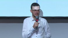 Eine Person im weißen Hemd spricht mit Mikrofon vor einem Bildschirm.