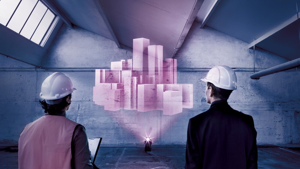 Zwei Ingenieure mit Schutzhelmen betrachten ein holografisches 3D-Gebäudemodell, das in eine Industriehalle projiziert wird.