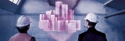 Zwei Personen, eine mit einer Warnweste und eine im Anzug und mit Helmen stehen in einer Halle und betrachten eine rosa Projektion von einem Gebäude im Raum.