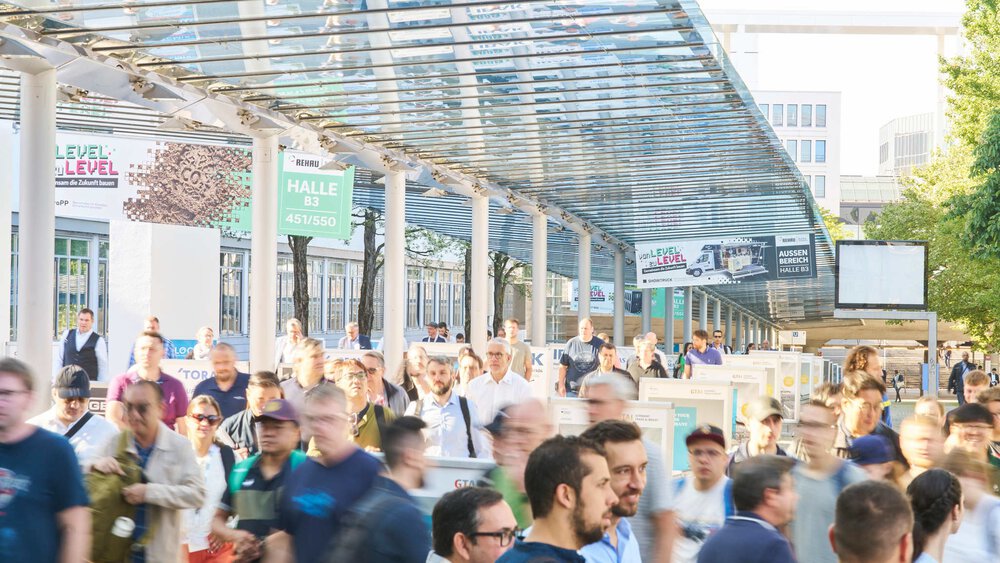 Messebesucher gehen unter einem überdachten Glasgang mit Wegweisern zu verschiedenen Hallen entlang des Außengeländes