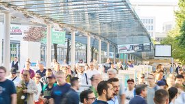 Messebesucher gehen unter einem überdachten Glasgang mit Wegweisern zu verschiedenen Hallen entlang des Außengeländes
