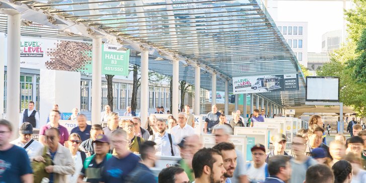 Messebesucher gehen unter einem überdachten Glasgang mit Wegweisern zu verschiedenen Hallen entlang des Außengeländes