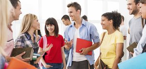 A group of smiling young adults chat in a bright interior with books and notebooks in their hands.