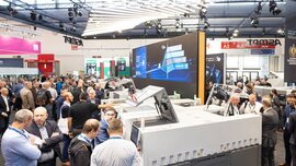 In einer Messehalle der productronica stehen zahlreiche Besucher um unterschiedliche, aufgeklappte Maschinen herum und unterhalten sich miteinander.