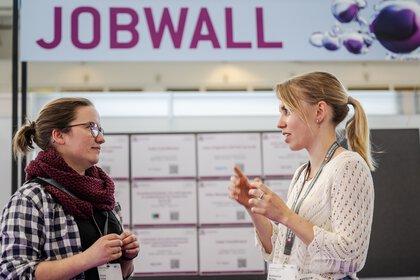 zwei Frauen stehen vor der sogenannten JOBWALL, einem Board mit Stellenanzeigen, und unterhalten sich